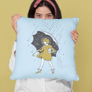 May include: A blue pillow with a girl holding an umbrella with a galaxy design. The girl is wearing a yellow dress and a helmet. The background is a light blue with stars and rain.