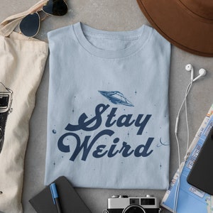 May include: A light blue t-shirt with a graphic of a UFO and the text "Stay Weird". The t-shirt is laid flat on a gray surface with a camera, a notebook, a tote bag, a hat, and headphones.