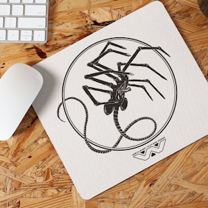 May include: A white mousepad featuring a black and white illustration of an alien creature within a circular frame. The mousepad also includes the text "WEYLAND-YUTANI CORP" and a logo. A white computer mouse and keyboard are visible.