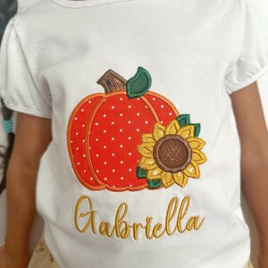May include: White t-shirt with an embroidered pumpkin with red and white polka dots and a sunflower. The name "Gabriella" is embroidered in gold below the pumpkin.