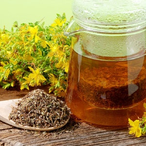 May include: A glass teapot filled with a golden yellow herbal tea, with dried St John's Wort herbs in a tea infuser. A wooden spoon holds loose St John's Wort herbs, and fresh St John's Wort flowers are scattered around the teapot.