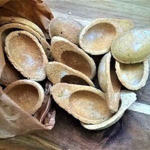 Op de afbeelding: Een bruine papieren zak gevuld met een grote hoeveelheid kleine, lichtbruine, komvormige gebakken producten. De producten zijn op een houten oppervlak in een stapel gelegd.