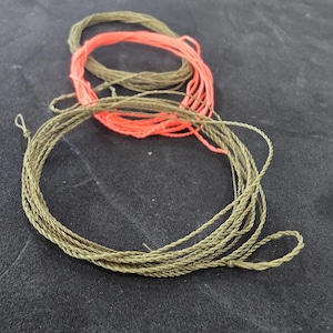 Puede incluir: Tres carretes de cuerda en tonos verde oliva y naranja coral. Las cuerdas están bien enrolladas y parecen estar hechas de un material de fibra trenzada. El fondo es una superficie oscura y texturizada.