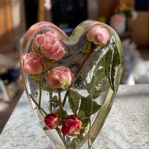 Puede incluir: Escultura de resina en forma de corazón con rosas rosas y rojas secas. La resina transparente encapsula las flores y las motas doradas. La escultura se apoya sobre una base transparente, creando una pieza decorativa única.