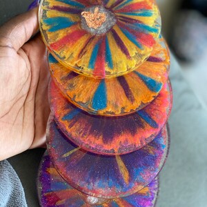 Puede incluir: Una pila de cinco posavasos de resina coloridos. Cada posavasos presenta un diseño único con líneas radiantes amarillas, naranjas, rojas, azules y moradas. Los posavasos tienen un acabado transparente y brillante y un toque de pan de oro en el centro.