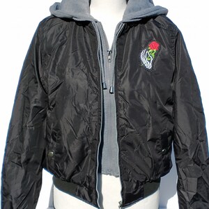Puede incluir: Chaqueta bomber negra con forro de capucha gris y una mano de esqueleto sosteniendo una rosa roja bordada en la parte delantera.