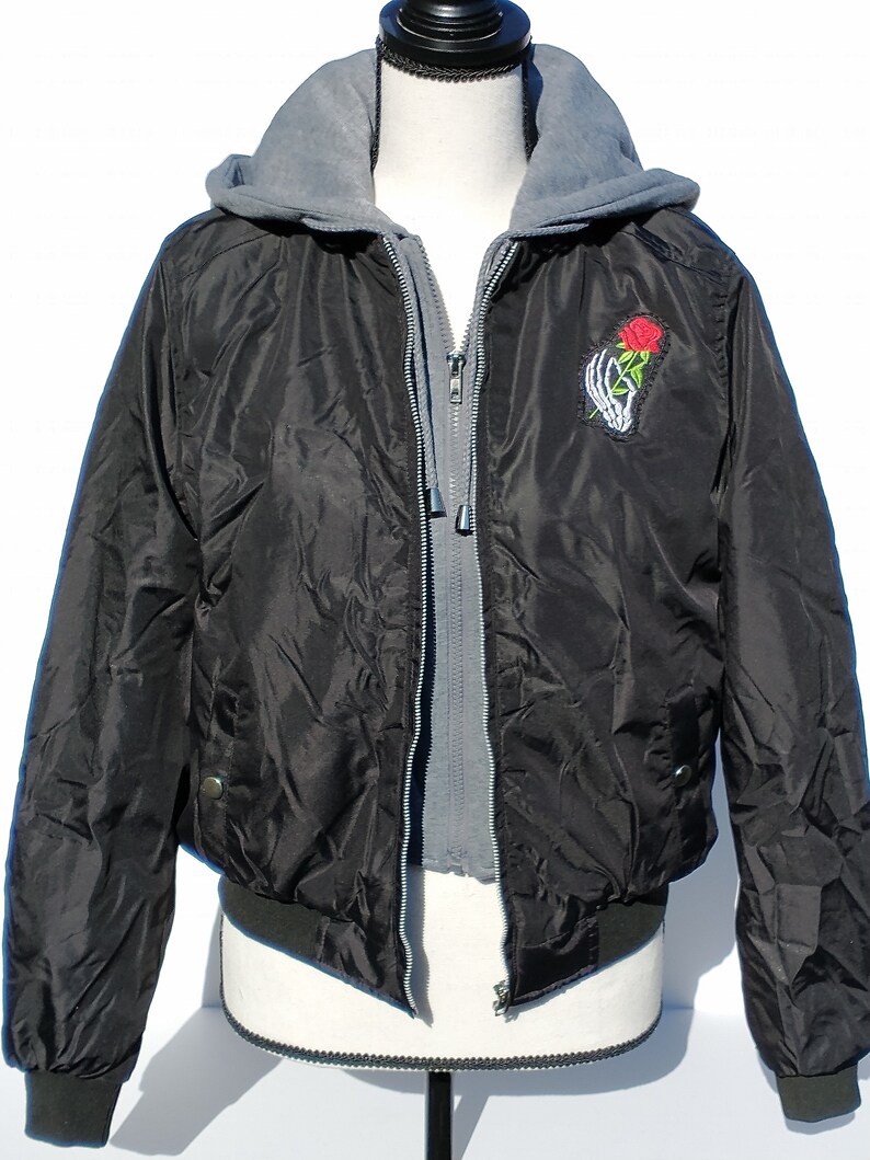 Puede incluir: Chaqueta bomber negra con forro de capucha gris y una mano de esqueleto sosteniendo una rosa roja bordada en la parte delantera.