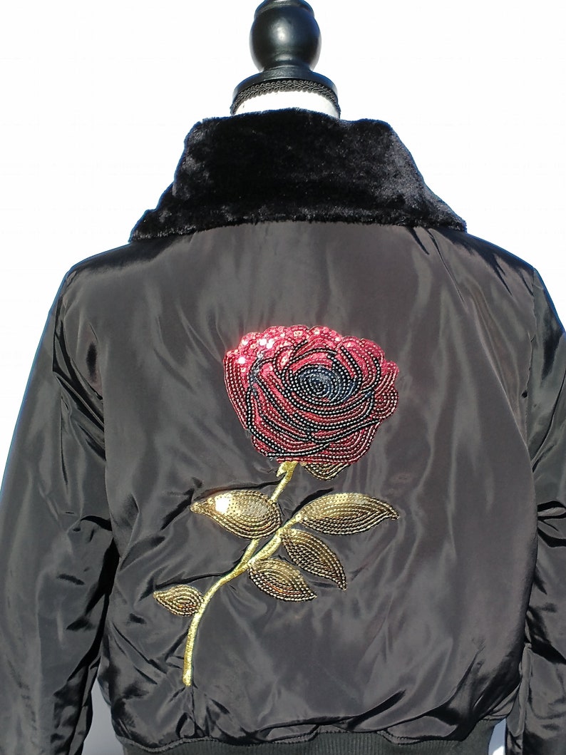 Puede incluir: Chaqueta bomber negra con cuello de piel sint&eacute;tica y un gran dise&ntilde;o de rosa de lentejuelas en la espalda.