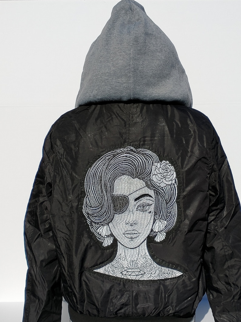 Puede incluir: Chaqueta bomber negra con una sudadera con capucha gris debajo. La chaqueta tiene un dise&ntilde;o bordado en la parte posterior de una mujer con un parche en el ojo y una flor en el pelo.