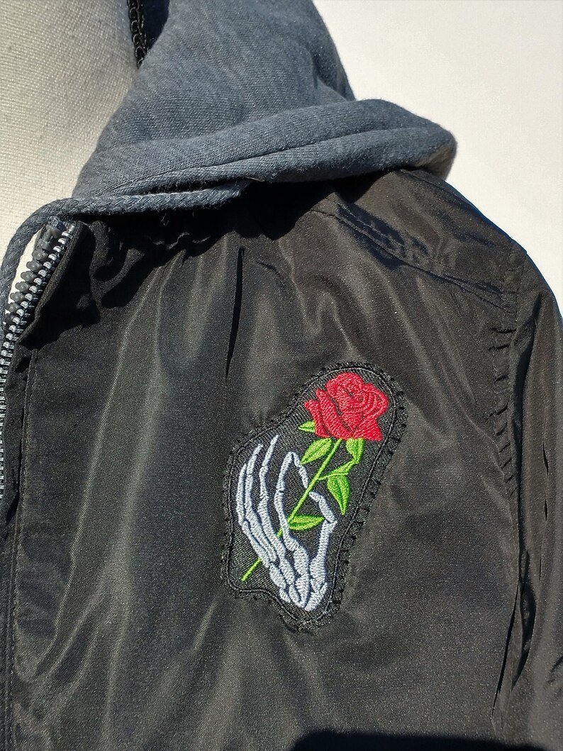 Puede incluir: Chaqueta bomber negra con una mano de esqueleto sosteniendo una rosa roja bordada en el lado izquierdo. La chaqueta tiene un forro de capucha gris.