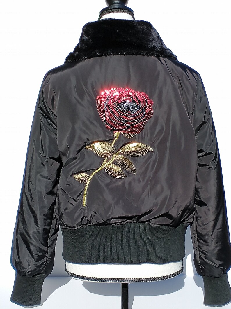 Puede incluir: Chaqueta bomber negra con cuello de piel sint&eacute;tica y un gran dise&ntilde;o de rosa de lentejuelas en la espalda.