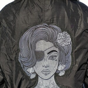 Puede incluir: Chaqueta bomber negra con una imagen bordada de una mujer con un parche en el ojo, una flor en el pelo y un tatuaje en el cuello.