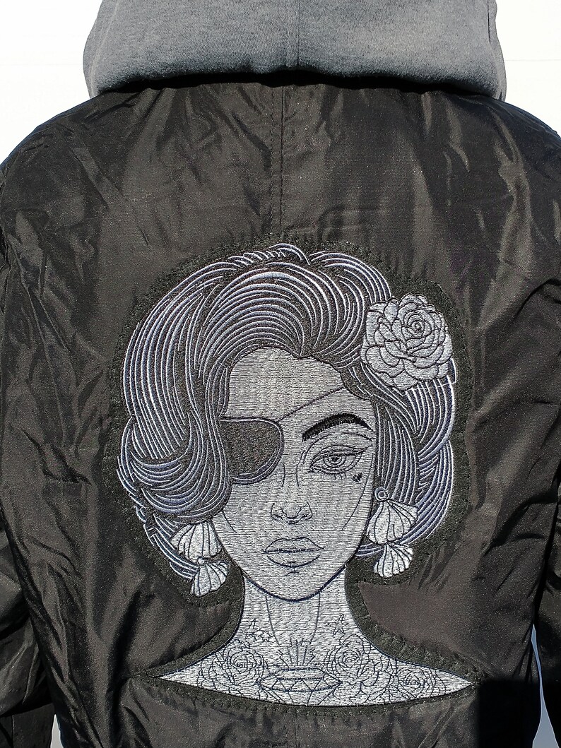 Puede incluir: Chaqueta bomber negra con una imagen bordada de una mujer con un parche en el ojo, una flor en el pelo y un tatuaje en el cuello.