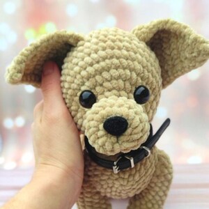 May include: A crocheted plush toy dog with brown fur, black eyes, and a black collar. The dog is being held by a hand.