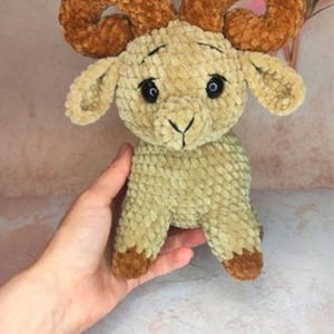 May include: A handmade plush ram toy with a light green body and brown horns and hooves. The ram has black eyes and a smiling expression. The toy is made of soft, textured fabric and is held by a hand.