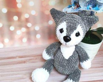 Può includere: Un giocattolo di peluche di lupo all'uncinetto grigio e bianco seduto su una superficie di legno. Il lupo ha una macchia bianca sul petto e piedi bianchi.