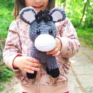 May include: A child is holding a grey and white crocheted donkey plush toy. The donkey has black eyes and a black mane. The child is wearing a pink floral jacket and pink trousers.