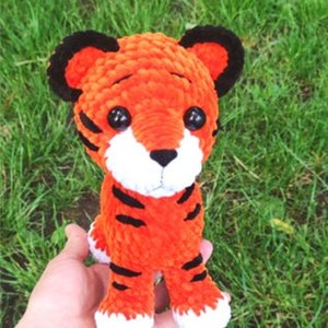 Puede incluir: Un juguete de peluche de tigre tejido a crochet con rayas naranjas y negras, patas blancas y ojos negros. El juguete está siendo sostenido en una mano contra un fondo de hierba verde.