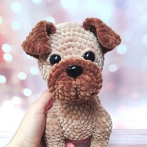 Puede incluir: Un perro de peluche tejido a crochet con orejas marrones y una cara marrón. El perro tiene ojos negros grandes y una nariz negra. El perro está sentado sobre una superficie marrón claro.