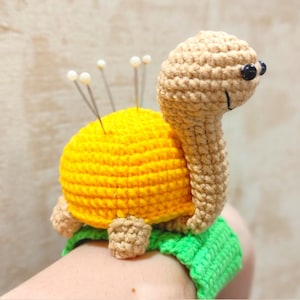 Puede incluir: Un cojín de alfileres de tortuga tejido a crochet con una concha amarilla y una cabeza y cuello marrones. La tortuga está sentada sobre una base verde tejida a crochet y tiene varios alfileres que sobresalen de su concha.