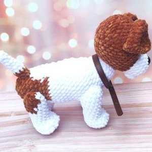 Op de afbeelding: Een wit en bruin gehaakt hondenspeelgoed met een bruin leren halsband. De hond staat op een houten oppervlak.