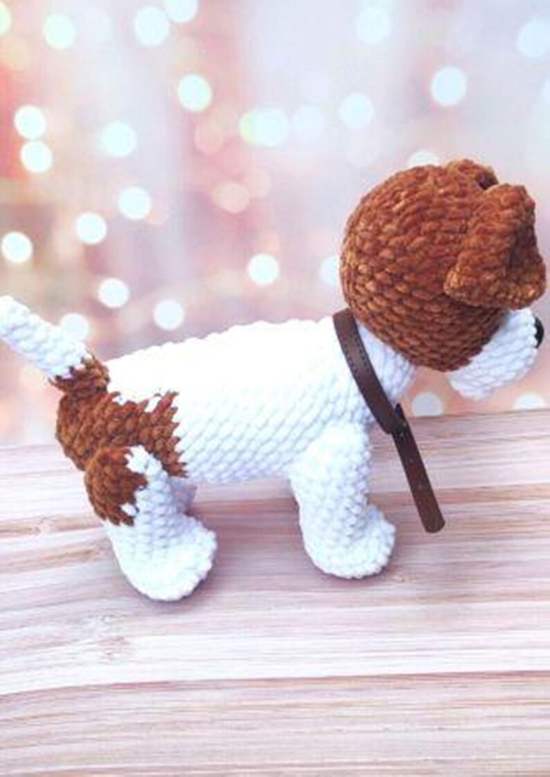 Op de afbeelding: Een wit en bruin gehaakt hondenspeelgoed met een bruin leren halsband. De hond staat op een houten oppervlak.