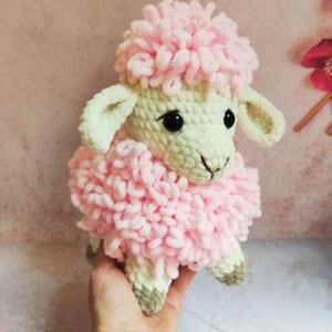 May include: A handmade crochet sheep toy with a fluffy pink head and body. The sheep has black eyes, light green ears, and light green feet with brown accents. The toy is held in a hand.