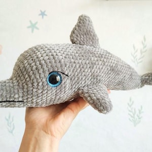 May include: A gray crocheted dolphin plush toy made from soft, textured yarn. The dolphin features a prominent dorsal fin, a tail, and large, round, bright blue plastic eyes with black pupils. The surface of the toy has a consistent, fuzzy, velvet-like appearance.