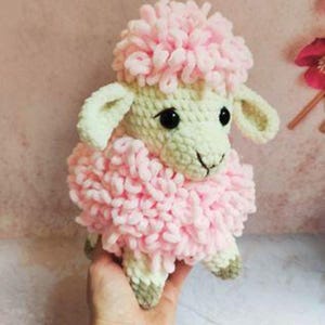 May include: A crocheted plush toy in the shape of a lamb. The lamb features a pale cream-colored face and legs with a textured, curly pink yarn body and head tuft. It has two small, dark, round eyes and a stitched nose and mouth detail.