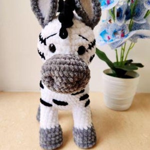 Może przedstawiać: Pluszowa zabawka w kształcie zebry wykonana na szydełku, z czarno-białymi paskami, szarymi uszami i kopytami. Zabawka stoi na drewnianej powierzchni przed doniczką z rośliną.