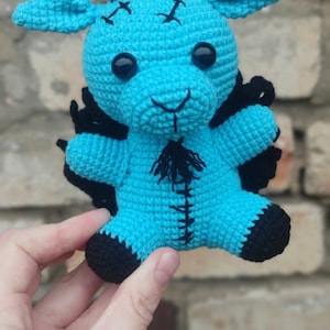 Könnte beinhalten: Ein helltürkisfarbenes Häkel-Plüschtier in Form einer Ziege mit schwarzen Hörnern, schwarzen Hufen und einem schwarzen Bart. Die Figur hat schwarze gestickte Symbole auf der Stirn, eine genähte Linie auf der Brust und schwarze Flügel am Rücken.