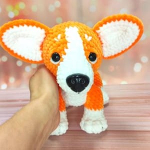 May include: A handmade orange and white crochet corgi dog plushie with large ears and a black nose. The plush toy is held by a hand and has a cute, friendly expression. The corgi is a popular dog breed.