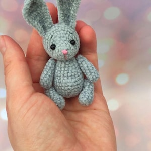 May include: A small, handmade, gray crochet bunny with long ears, black eyes, and a pink nose. The bunny is held in a hand, showcasing its soft texture and detailed craftsmanship. A charming, miniature stuffed animal.