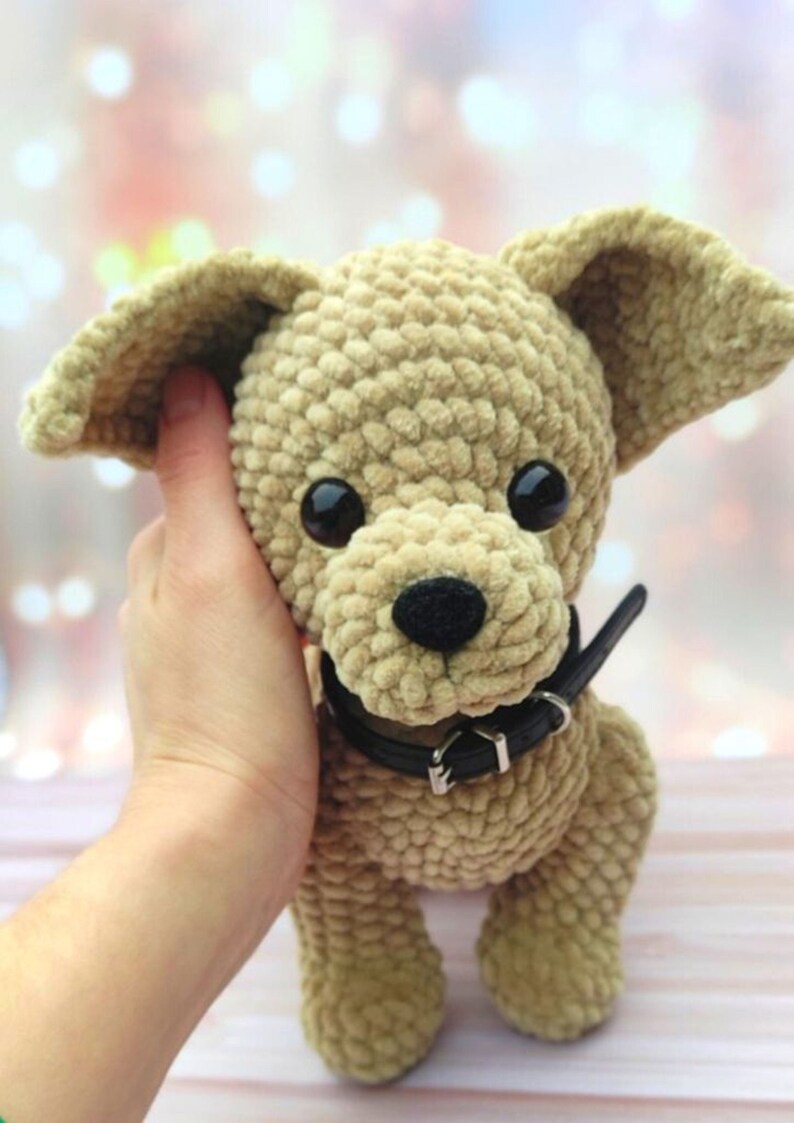 May include: A crocheted plush toy dog with brown fur, black eyes, and a black collar. The dog is being held by a hand.