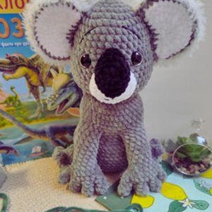 May include: A handmade gray crochet koala bear with large white-edged ears, a black nose, and round black eyes. The koala is sitting upright. A colorful book with dinosaur illustrations is in the background.