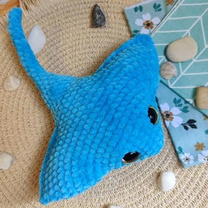 May include: A handmade, plush blue stingray toy with black eyes and a textured surface. The toy is lying on a woven straw mat, surrounded by seashells and decorative elements. A whimsical, ocean-themed item.