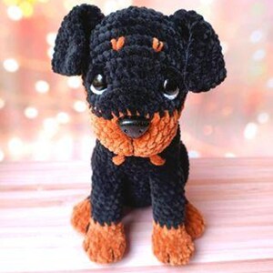 May include: A crocheted plush toy Rottweiler dog with black fur and brown accents. The dog is sitting with its paws tucked under its body and has a sweet expression on its face.
