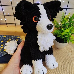 Puede incluir: Un perro de ganchillo hecho a mano en blanco y negro, con ojos naranjas y una nariz negra. El peluche tiene patas blancas y un pecho blanco y esponjoso. El perro est&aacute; sentado sobre una estera tejida.
