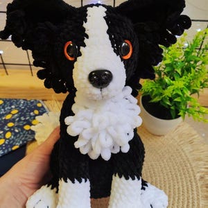 以下が含まれることがあります： オレンジ色の目と白いふわふわの襟が付いた、手作りの白黒のぬいぐるみ犬。犬は織りのマットの上に座っています。犬の耳は黒くて垂れ下がっており、足は白です。