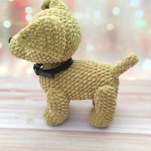 May include: A plush tan-colored dog toy with a black collar. The toy is standing on a light wood surface.