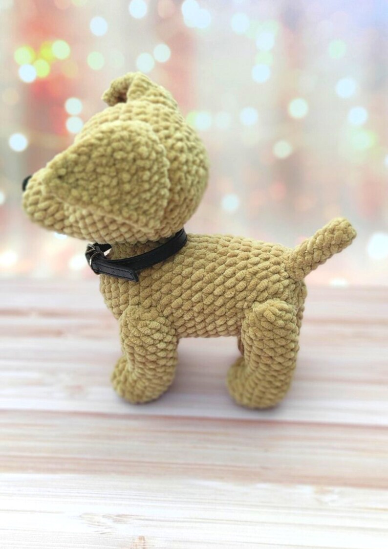 May include: A plush tan-colored dog toy with a black collar. The toy is standing on a light wood surface.