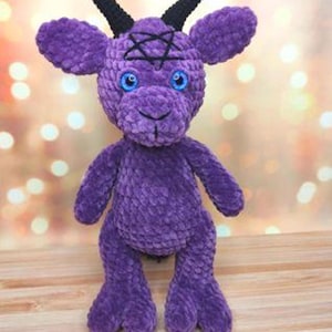 May include: A plush, purple crochet Baphomet doll with black horns and a black pentagram on its forehead. The doll has blue eyes and is standing on a wooden surface. The plushie is a unique handmade item.