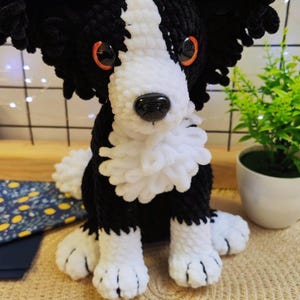 Puede incluir: Un perro de ganchillo hecho a mano en blanco y negro, con ojos naranjas y una nariz negra. El perro tiene un pecho y patas blancos y esponjosos. El perro est&aacute; sentado sobre una estera tejida.