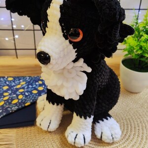 Puede incluir: Un perro de crochet hecho a mano en blanco y negro, con ojos grandes y expresivos. El peluche est&aacute; sentado sobre una estera tejida, mostrando su detallada artesan&iacute;a. El perro tiene pecho, patas y cara blancos, con detalles negros.