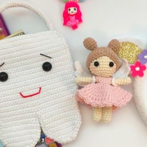 May include: A white crocheted tooth-shaped bag with a smiling face, a crocheted fairy doll in a pink dress, and a pink beaded necklace with a doll pendant. The items are arranged on a white surface.