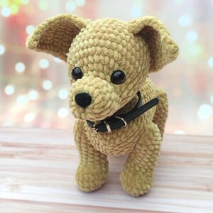 May include: A crocheted plush toy dog with brown fur, black eyes, and a black leather collar. The dog is sitting on a light brown wooden surface.