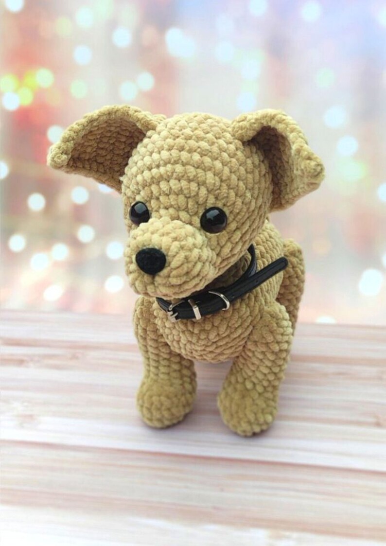 May include: A crocheted plush toy dog with brown fur, black eyes, and a black leather collar. The dog is sitting on a light brown wooden surface.