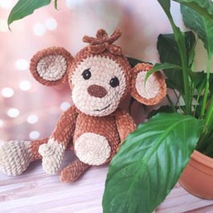 May include: A brown and cream crocheted monkey plush toy with a smiling face. The monkey is sitting on a light wood surface with a green plant in the background.