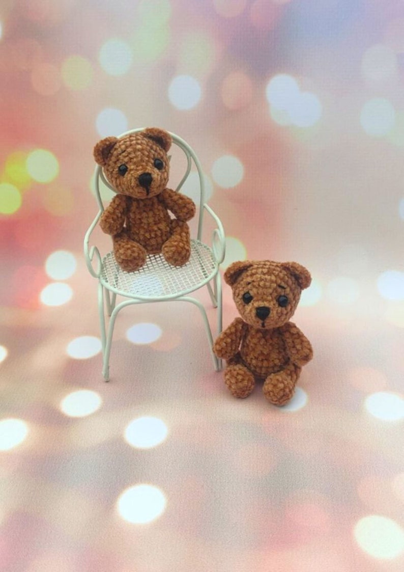 May include: Two handmade brown crochet teddy bears. One teddy bear is sitting on a small white chair, and the other is sitting on the surface. The bears have black eyes and noses. The background is a soft pink with bokeh lights.