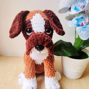 Puede incluir: Un perro de peluche de crochet marrón y blanco con ojos grandes. El perro está sentado con las patas metidas debajo del cuerpo.